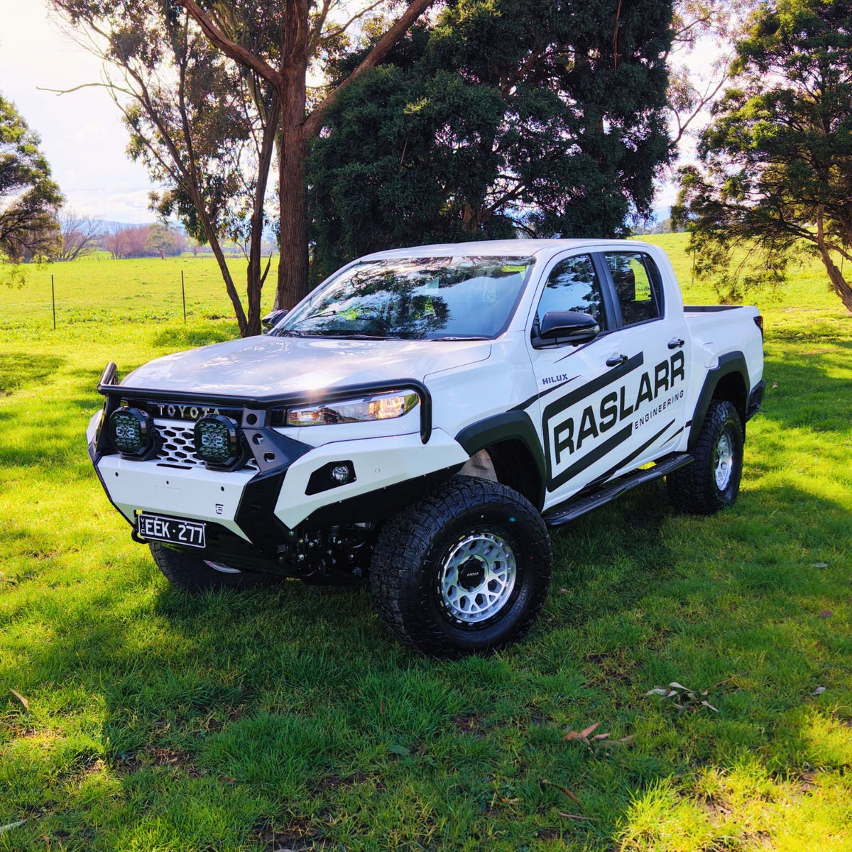 A white Toyota Hilux pickup truck with custom black graphics and off-road modifications, parked on a grassy lawn with trees in the background.
